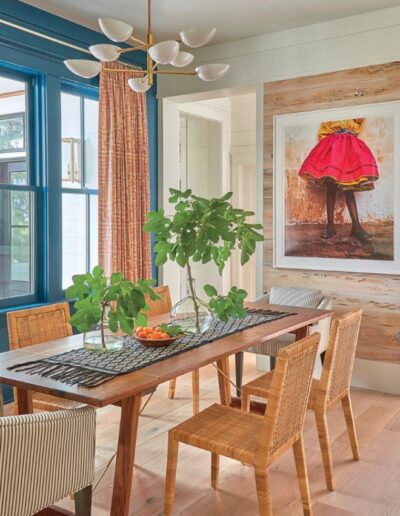 Dining room in the 2018 Coastal Living Idea House with a wood table, woven chairs, a brass chandelier, and blue window trim.