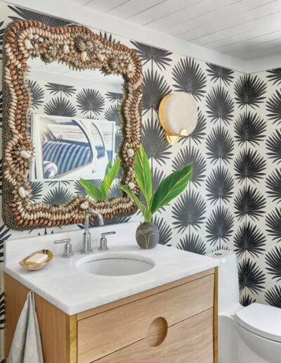Bathroom in the 2018 Coastal Living Idea House with a wood vanity, marble top, shell-framed mirror, and palm-patterned wallpaper.