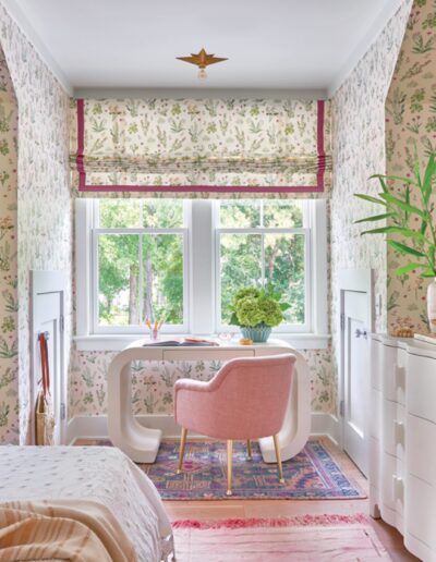 Bedroom in the 2018 Coastal Living Idea House with floral wallpaper, a curved desk, a pink chair, and a window overlooking trees.