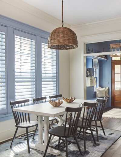 Dining area with a white trestle table, black Windsor chairs, and a woven rattan dome pendant beneath blue plantation shutters in the 2020 Southern Living Showcase Home in Habersham, Beaufort, SC.