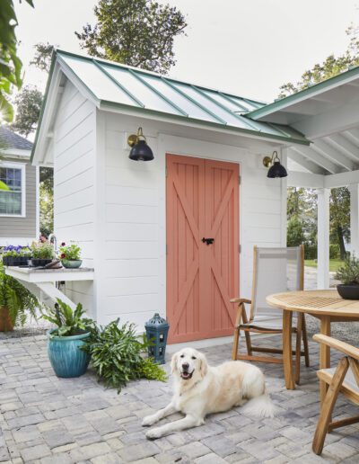 Garden shed with a coral barn door and green standing-seam metal roof at the 2020 Southern Living Showcase Home in Habersham, Beaufort, SC, with an attached potting bench and a golden retriever resting on the paver pati